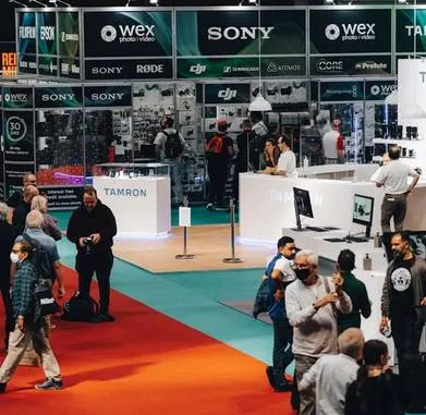 People walking and interacting at a busy technology or camera trade show with booths for brands like Sony, Tamron, and Wex Photo Video