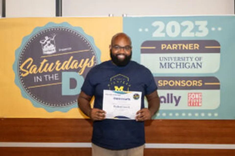 Rapheal French holding his certificate in front of a sign promoting Saturdays in the D